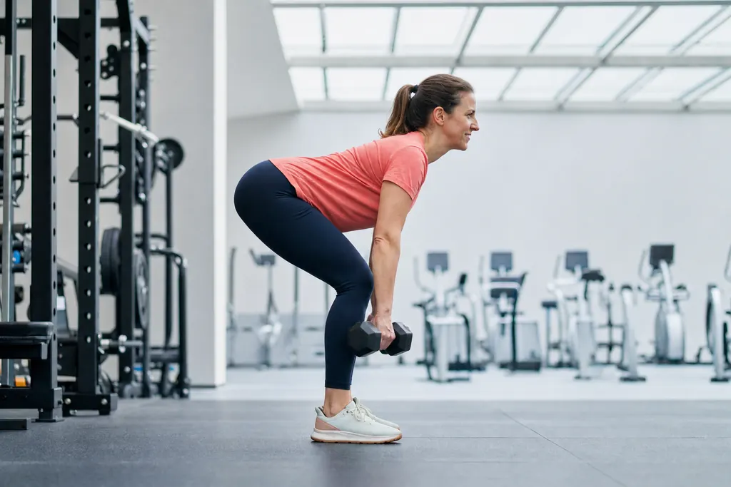 Woman doing a dumbbell Romanian deadlift in a bright gym, side view